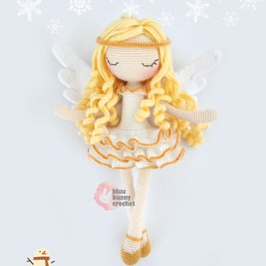 May include: A crocheted angel doll with blonde hair, white wings, and a white dress with gold trim. The doll is standing on a snowy surface with snowflakes in the background. The text "bluu bunny crochet" is visible in the image.
