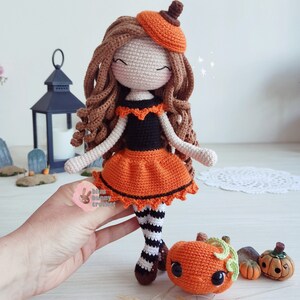May include: A crocheted doll wearing an orange and black dress and a pumpkin hat. The doll is holding a small crocheted pumpkin. The doll is standing on a wooden surface.