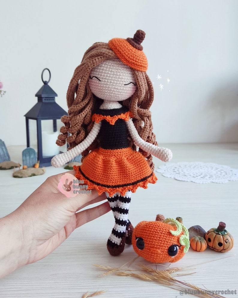 May include: A crocheted doll wearing an orange and black dress and a pumpkin hat. The doll is holding a small crocheted pumpkin. The doll is standing on a wooden surface.