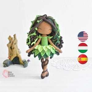 Puede incluir: Muñeca de ganchillo hecha a mano con piel morena, vestida con un vestido y pelo verdes con forma de hoja. El pelo de la muñeca es una mezcla de hilo marrón y verde, peinado con detalles de hojas. La muñeca está sobre una superficie blanca.