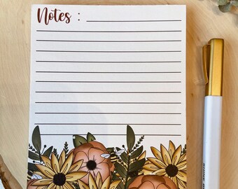 May include: A rectangular notepad with horizontal lines and the word Notes printed at the top. The bottom features a floral illustration of yellow sunflowers, orange flowers, green leaves, and a small bee. A white pen with a gold cap is beside it.