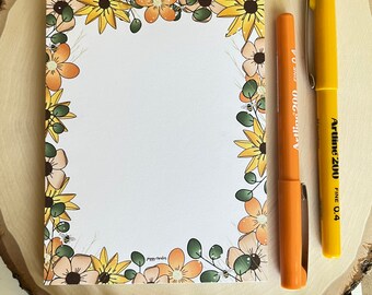 May include: A rectangular notepad with a white center and a border of illustrated yellow and orange flowers, green leaves, and small bees. The text "piggy creates" is printed at the bottom. An orange marker and a yellow marker are placed to the right.