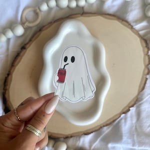 May include: A die-cut sticker featuring a white ghost with black oval eyes, holding a red soda can with a black straw. The ghost has a simple, cartoon-style outline with subtle gray shading to define the folds of the sheet-like body.