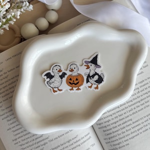 May include: A white, wavy-edged ceramic dish holds three Halloween-themed duck stickers. One duck has bat wings, another is a jack-o'-lantern, and the third wears a witch hat. The stickers are on top of an open book.