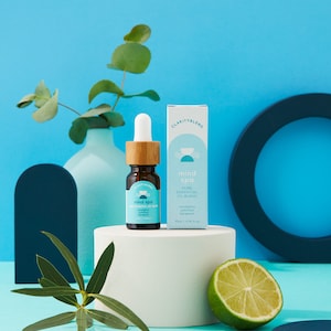 May include: A brown glass bottle with a dropper and a wooden cap, labelled "mind spa" with a light blue and white design. A white box with the same design and text is next to the bottle. A lime and eucalyptus sprig are in the foreground.