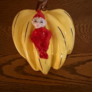 May include: A ceramic pumpkin-shaped decoration with a smiling elf figurine. The pumpkin is yellow with brown accents and vertical lines. The elf wears a red outfit and hat. A whimsical autumn-themed decorative item.