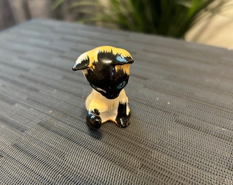May include: A small ceramic figurine of a black and white cat sitting with its head down. The cat has blue eyes and is painted in a realistic style.