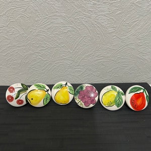 May include: Six ceramic knobs with fruit designs. The knobs are white with painted images of cherries, pears, grapes, lemons, and a red pear.