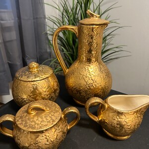 May include: A gold-colored ceramic tea set with a teapot, creamer, and sugar bowl. The set has a textured, embossed design.