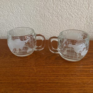 May include: Two clear glass mugs with handles. Each mug features a world map design etched onto the glass. The mugs are sitting on a wooden surface.