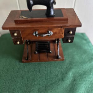 May include: Miniature wooden sewing machine with a black machine head. The sewing machine has a brown wooden table with a drawer and two small side drawers. The machine is set on a green cloth.
