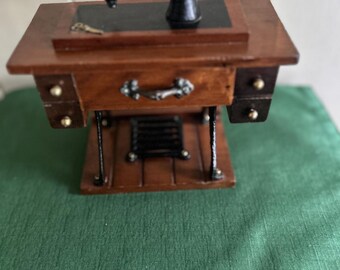 May include: Miniature wooden sewing machine with a black machine head. The sewing machine has a brown wooden table with a drawer and two small side drawers. The machine is set on a green cloth.