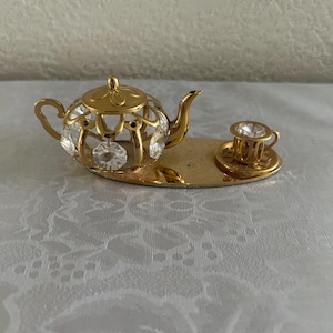 May include: A gold-tone tea set figurine with a teapot and cup on a tray. The teapot and cup are decorated with clear crystals.