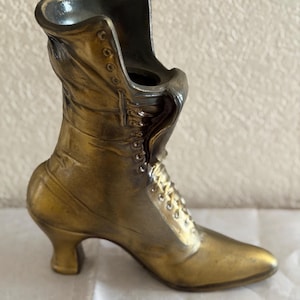 May include: A bronze-colored decorative boot with a pointed toe and a small heel. The boot features a lace-up design and is likely a vintage or antique item. The boot is made of metal and has a glossy finish.