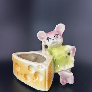 May include: A whimsical ceramic planter featuring a mouse figure leaning against a cheese wedge. The mouse has pink ears and a green outfit. The cheese wedge is yellow with round holes, creating a charming, vintage-inspired decorative piece.