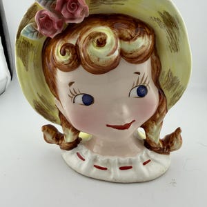 May include: A vintage ceramic head vase featuring a girl with brown curly hair, blue eyes, and a red mouth. She wears a yellow hat adorned with pink roses and green leaves. The vase has a white collar with red accents.