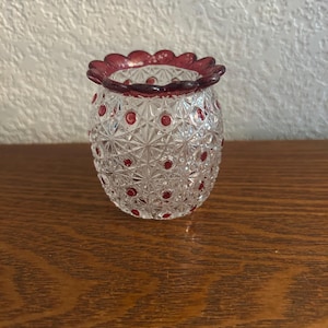May include: A clear glass vase with a cut crystal pattern and red accents. The rim of the vase is a deep red with a scalloped edge. Small red dots are evenly spaced across the body of the vase. The vase is sitting on a wooden surface.