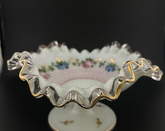 May include: A white footed bowl with a ruffled edge and gold trim. The bowl is decorated with pink and blue flowers and gold accents.