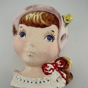 May include: Vintage ceramic wall pocket in the shape of a girl's head. The girl has blue eyes, brown hair, and a pink bonnet. A yellow flower accents the bonnet. A red bow adorns the white collar with blue polka dots. A charming decorative piece.