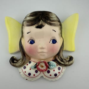 May include: A vintage ceramic wall hanging featuring a girl's face with blue eyes, rosy cheeks, and brown hair. The girl has yellow wings and a white collar with purple polka dots. A red and white flower is on the collar.