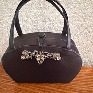 May include: A dark brown vintage handbag with a decorative silver-toned floral embellishment. The bag has a rounded shape, a top handle, and a button closure. The bag is sitting on a wooden surface.