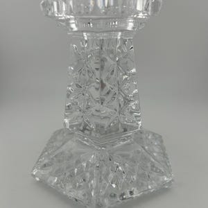 May include: A clear glass candle holder with a geometric design. The holder has a wide base and a tall, slender column.