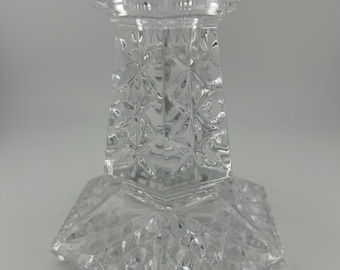 May include: A clear glass candle holder with a geometric design. The holder has a wide base and a tall, slender column.