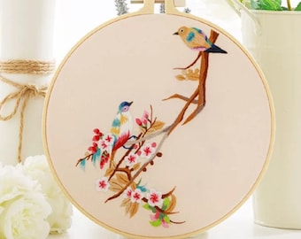 May include: A round embroidery hoop featuring a detailed hand-stitched design. Two colorful birds perch on a brown branch with pink and white flowers. The fabric is a light beige color, and the hoop is made of natural wood.