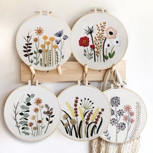 May include: Four embroidery hoops with floral designs. The hoops are hanging on a wooden wall rack. The designs include a variety of flowers, leaves, and stems in shades of red, yellow, green, and brown.