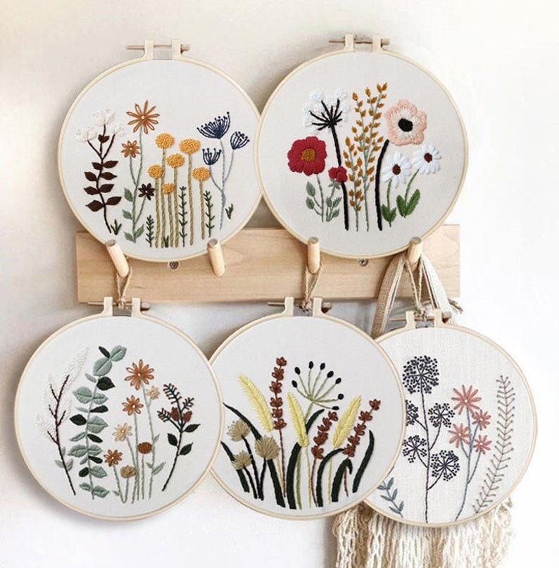 May include: Four embroidery hoops with floral designs. The hoops are hanging on a wooden wall rack. The designs include a variety of flowers, leaves, and stems in shades of red, yellow, green, and brown.