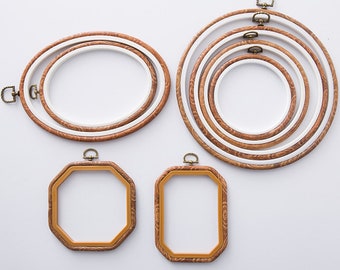 Puede incluir: Un conjunto de bastidores de bordado de varias formas y tamaños. Los bastidores presentan un patrón de vetas de madera en el anillo exterior y un anillo interior blanco. El conjunto incluye marcos ovalados, redondos, octogonales y rectangulares, todos con un cierre de color latón.