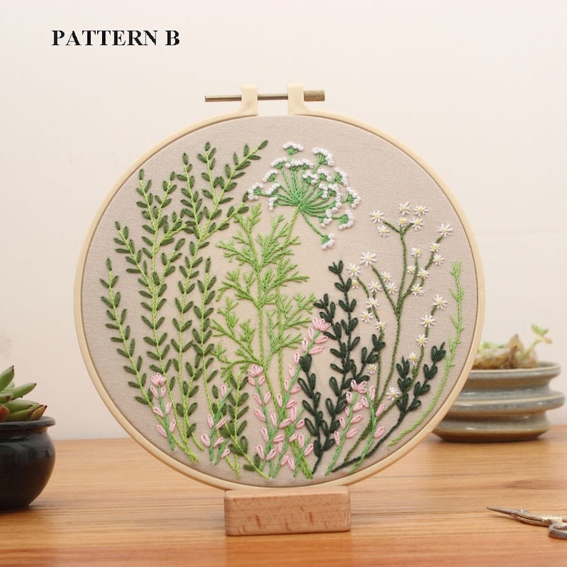 May include: A round embroidery hoop with a beige fabric background featuring a floral design. The design includes green leaves, pink flowers, and white flowers. The hoop is made of wood and has a natural finish.