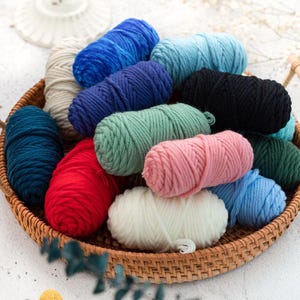 May include: A wicker basket filled with various colorful yarn skeins. The yarn is in shades of blue, green, pink, red, black, and cream. The yarn is arranged in a visually appealing way, suggesting a craft or hobby theme.