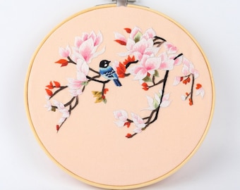 May include: Embroidered floral hoop art featuring a branch with pink magnolia blossoms and a blue bird. The embroidery is set against a peach-colored fabric, framed by a wooden hoop. The bird has blue and black details.