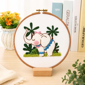 Peut inclure: Un tambour à broder circulaire en bois contenant un tissu blanc avec un motif brodé à la main représentant un éléphant blanc avec un nœud rose, des détails bleus et des palmiers verts. Le tambour est posé sur un support en bois rectangulaire.