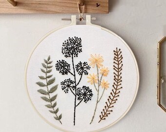 May include: A round embroidery hoop with a white fabric featuring embroidered floral designs. The design includes green fern-like leaves, black seed heads, and yellow and brown flowers. The hoop is hanging on a wooden wall.