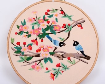 May include: Embroidered artwork featuring two blue birds perched on a branch with pink blossoms and red berries. The design is set within a round wooden embroidery hoop, showcasing a nature-themed scene.