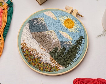 Puede incluir: Arte de bordado en aro que presenta una escena de paisaje con montañas, árboles y un sol. Las montañas son una mezcla de azul, gris y marrón, y los árboles son verdes. El sol es amarillo y el cielo es azul con nubes blancas.