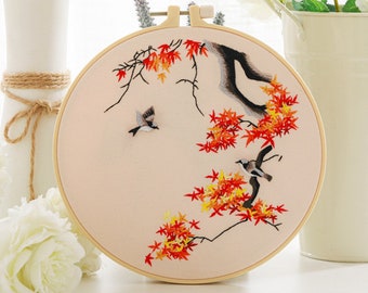 May include: A circular wooden embroidery hoop containing a light beige fabric panel. The fabric features detailed needlework of a dark tree branch with vibrant red and yellow maple leaves, accompanied by two small birds in flight and perched.