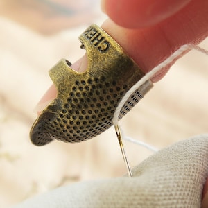 Peut inclure: Un dé à coudre en métal doré avec une surface texturée et le mot "CHEF" en relief sur le côté. Le dé à coudre est utilisé pour enfiler une aiguille avec du fil blanc.