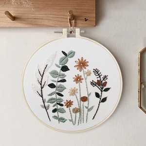 May include: A white embroidery hoop with a floral design. The design features green leaves, white flowers, and orange and brown flowers.