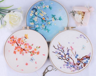 May include: Three embroidery hoops with floral designs. The hoops are made of wood and have a white fabric stretched over them. The designs are of cherry blossoms, with pink, purple, and blue flowers. One hoop features a bluebird on a branch.