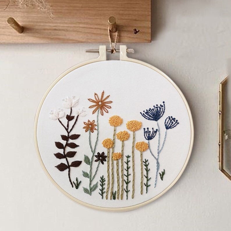 May include: Embroidered hoop art featuring a floral design with white, brown, yellow, blue, and green flowers. The hoop is hanging on a wooden wall.