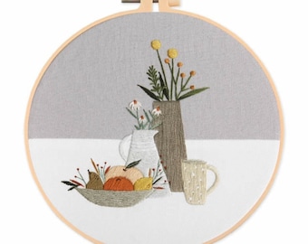 Puede incluir: Arte de bordado en aro que representa una naturaleza muerta con un jarrón de flores, una jarra, un cuenco de fruta y una taza. El fondo es gris claro y el primer plano es blanco.
