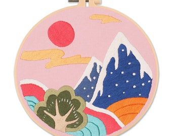 May include: Embroidered landscape art in a wooden hoop. The design features a red sun, blue mountains with white snow, a green tree, and colorful ground details against a pink sky. The embroidery is secured in a round wooden hoop.