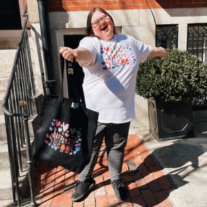 May include: A person wearing a white t-shirt and gray sweatpants, holding a black tote bag. The t-shirt and tote bag both feature the text "DIFFERENT IS COOL" surrounded by colorful hearts. The person is smiling and standing outside.