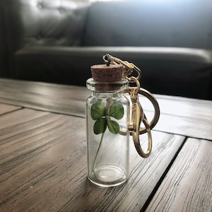 May include: A small glass bottle with a cork top and a gold chain attached. Inside the bottle is a preserved four-leaf clover.