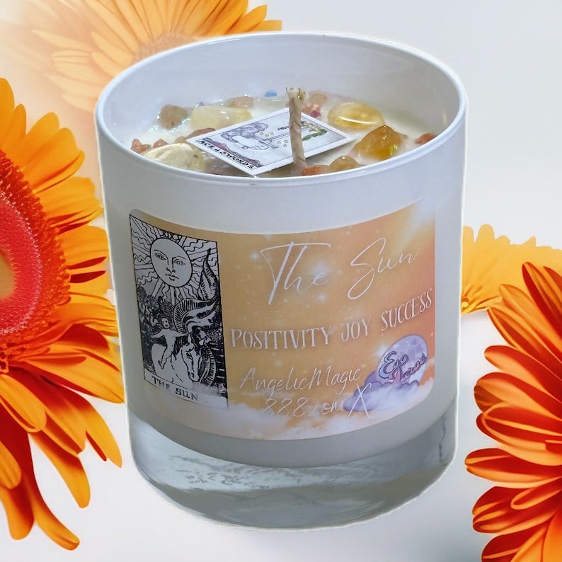Op de afbeelding: Witte kaars met een geel en oranje etiket met de tekst "The Sun" met de tekst "Positivity Joy Success" en "Angelic Magic 228 cm". De kaars is omringd door oranje bloemen.