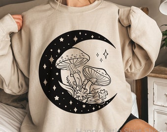 May include: A beige sweatshirt with a black crescent moon graphic featuring two mushrooms and stars. The text "Happy Little Shirt Shop" is visible at the bottom of the image.