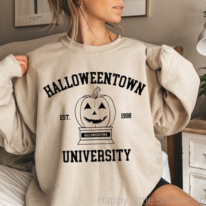 Puede incluir: Una sudadera beige con un estampado gr&aacute;fico negro de una calabaza con una cara sonriente. El texto "HALLOWEENTOWN UNIVERSITY EST. 1998" est&aacute; impreso en la sudadera.